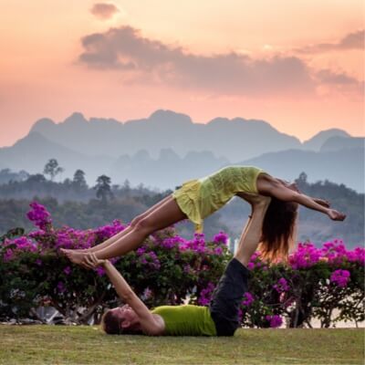 A man lyning on the ground balancing a woman in a yoga pose set against a sunset.